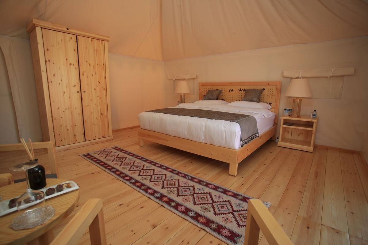 Een tentkamer met een houten bed, twee nachtkastjes, een houten kledingkast en een kleine tafel met drankjes en snacks.