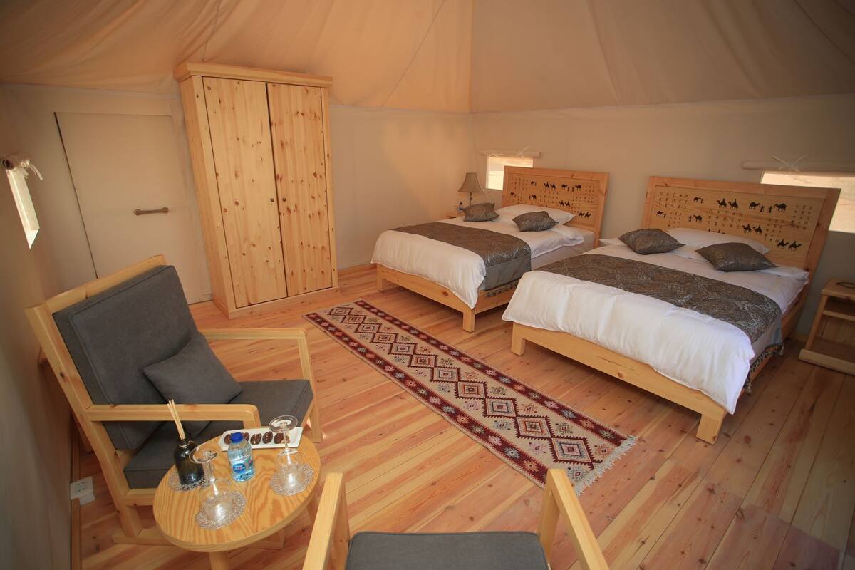 Een tentaccomodatie met twee houten bedden, een kledingkast, een zithoek met een tafel en stoelen, en een vloerkleed met een patroon.