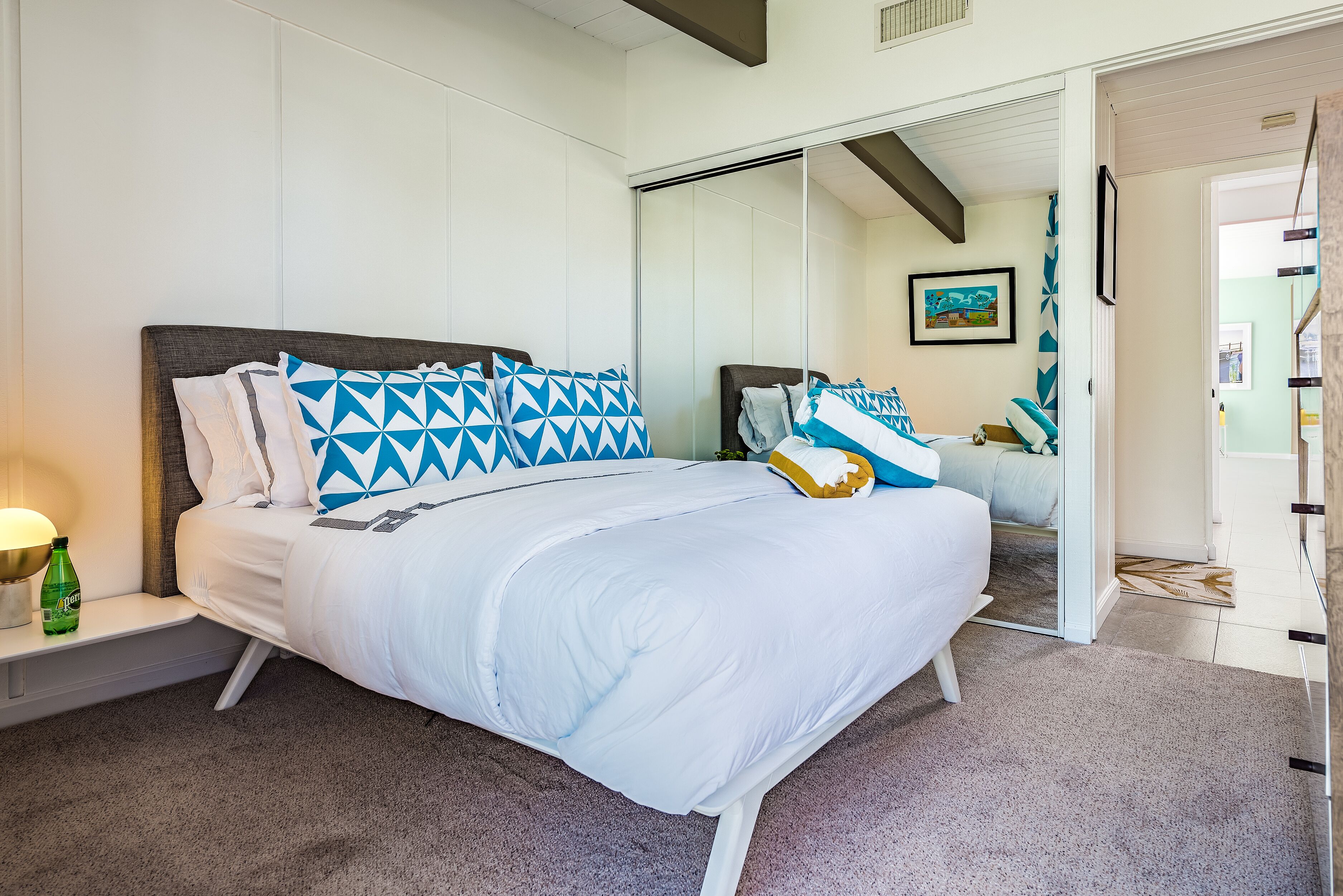 A neatly made bed with white bedding and blue geometric pillows, a bedside table with a green bottle, and a large mirror reflecting the room.