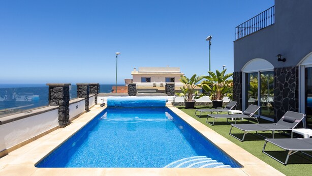 A swimming pool with a clear view of the ocean, surrounded by lounge chairs and a building with a stone facade. - Villa Syrah Tenerife (Puerto De La Cruz)