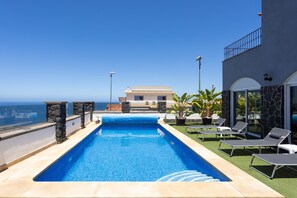 A swimming pool with a clear view of the ocean, surrounded by lounge chairs and a building with a stone facade.
