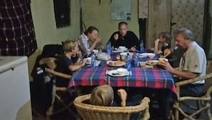 A group of people dining at a table with a plaid tablecloth. The room has a rustic appearance with a thatched roof and wicker chairs. - Leruk Guest House (Maasai Mara)