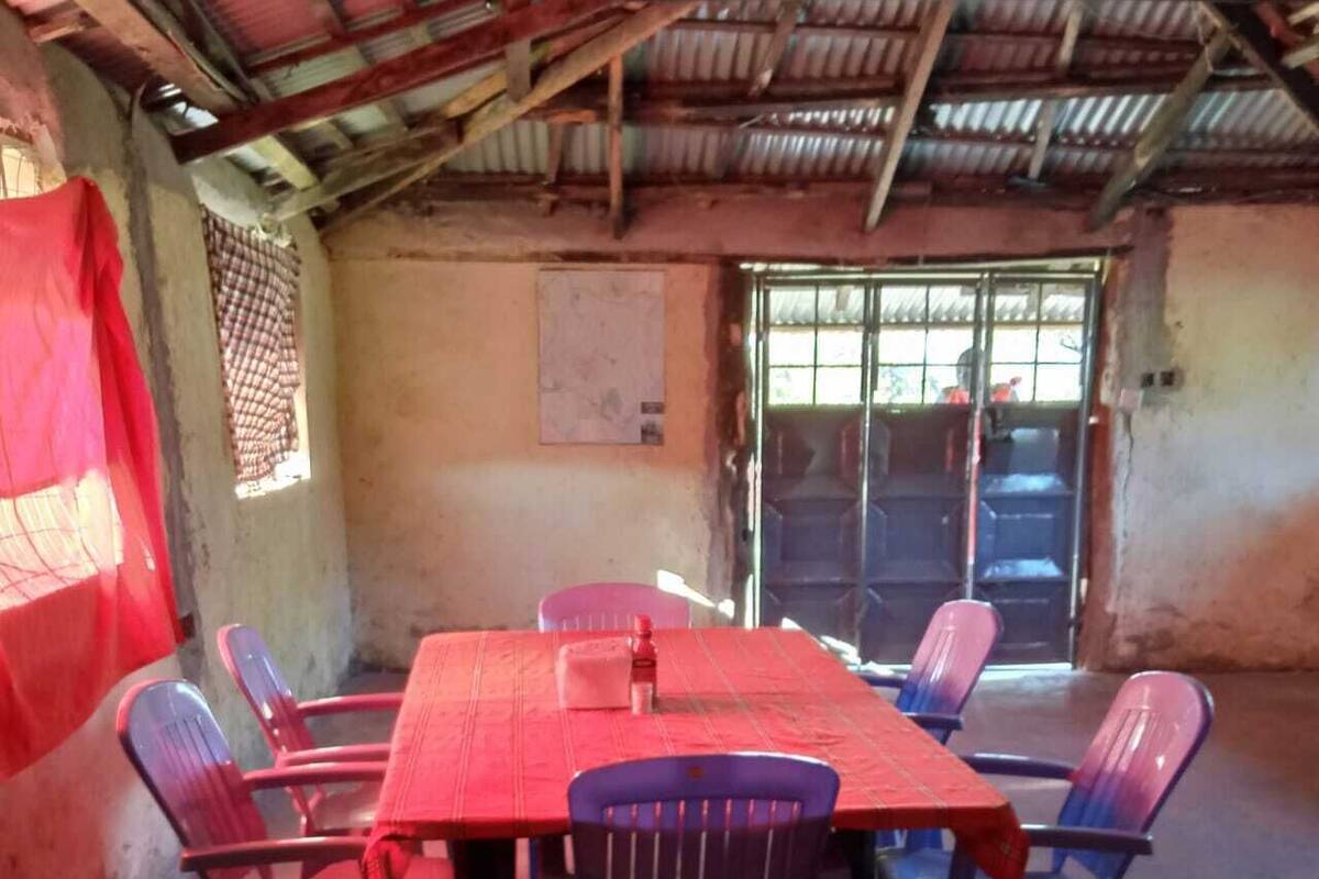A room with a red table and plastic chairs, a window with bars, and a door leading outside.