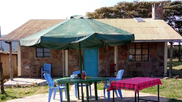 A stone building with a tiled roof, a covered patio with a table and chairs, and a red checkered tablecloth on the ground. - Leruk Guest House (Maasai Mara)