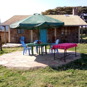 Um edifício de pedra com telhado ladrilhado, um alpendre coberto com mesa e cadeiras, e uma toalha xadrez vermelha no chão.