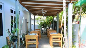 Un patio al aire libre con mesas y sillas de madera, rodeado de macetas con plantas y árboles.