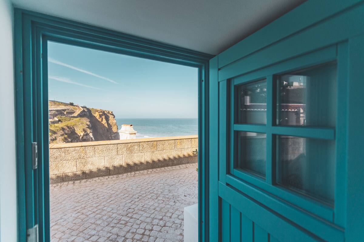 A view from a room with a teal door and window, overlooking a cobblestone path leading to a cliffside and the ocean.