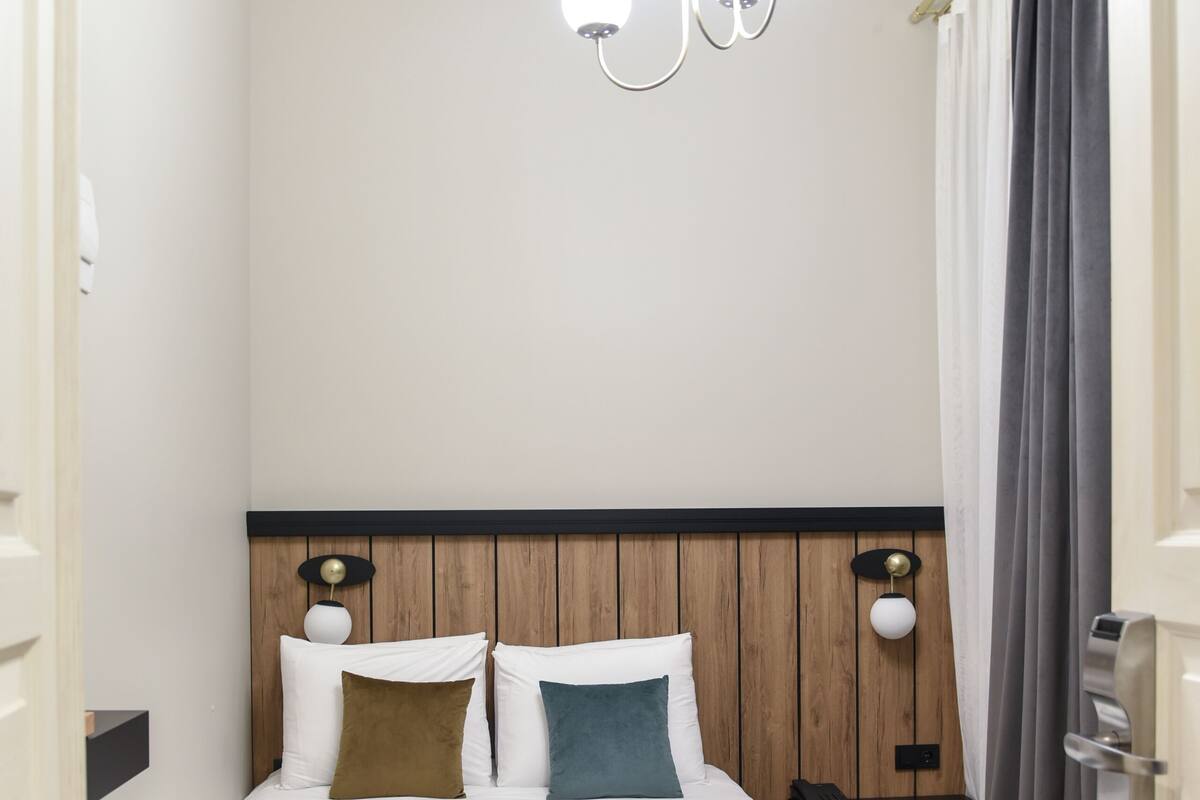 A modern hotel room with a bed, a wooden headboard, a nightstand, a lamp, and a hanging light fixture.