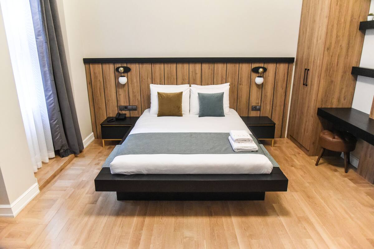 A modern hotel room with a large bed, wooden headboard, bedside tables with lamps, and a small desk with a chair.