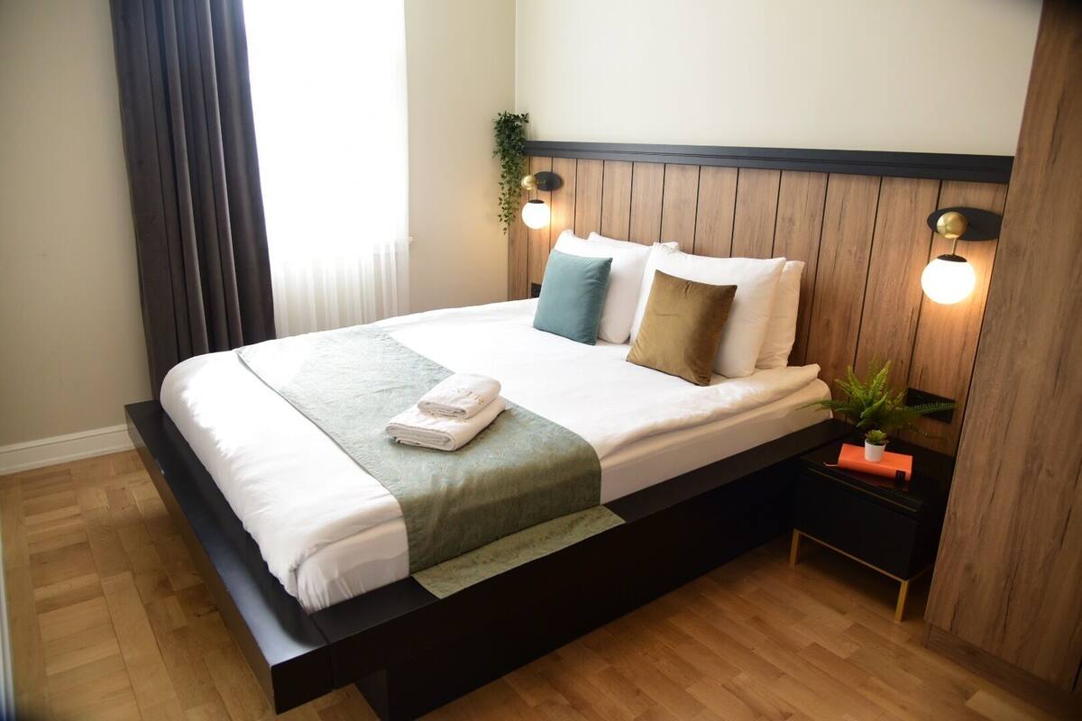A neatly made bed with a white comforter and multiple pillows, a dark wooden headboard, and two wall-mounted lights.
