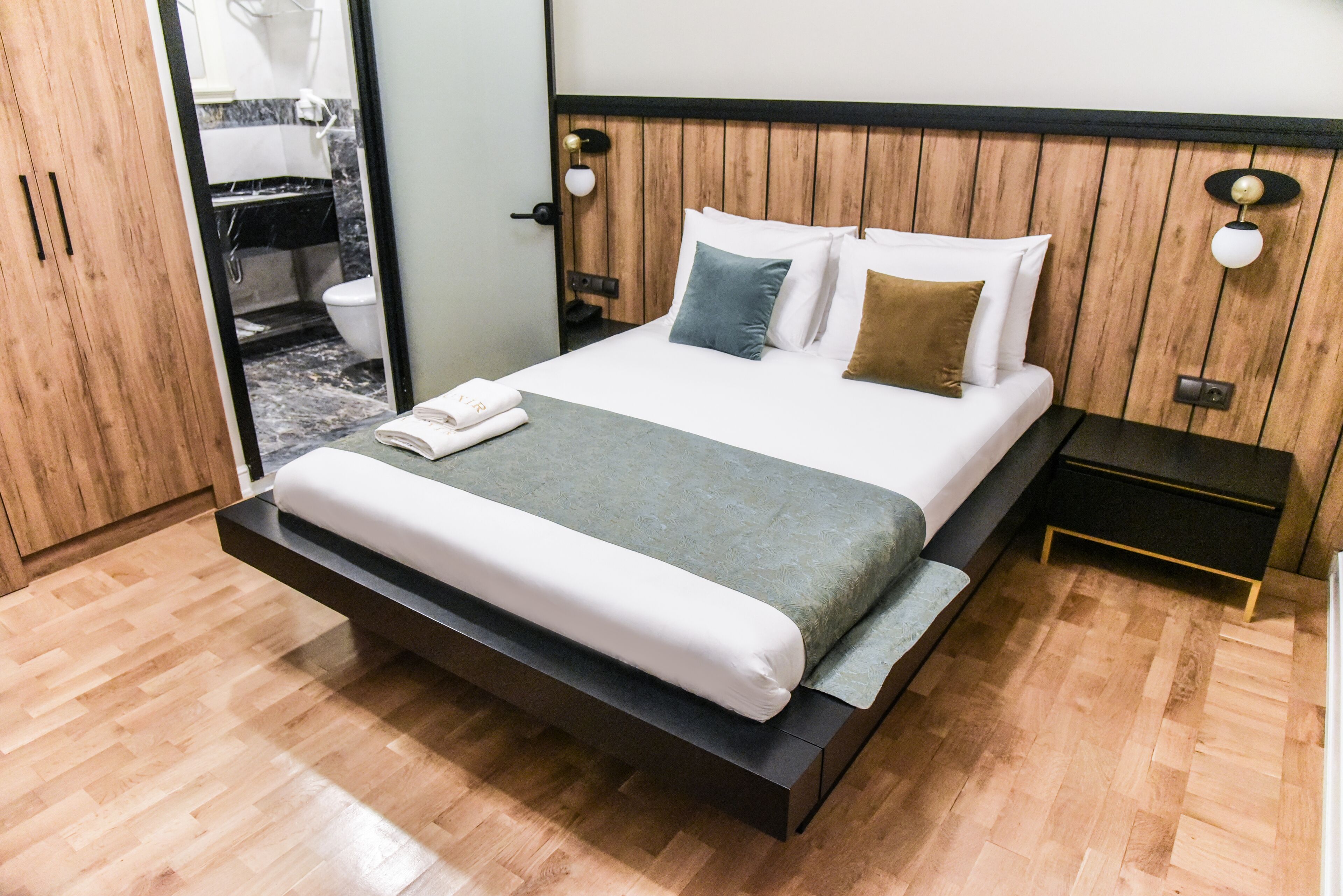 A modern bedroom with a wooden floor, a bed with white and grey bedding, a nightstand, and a wooden wardrobe.