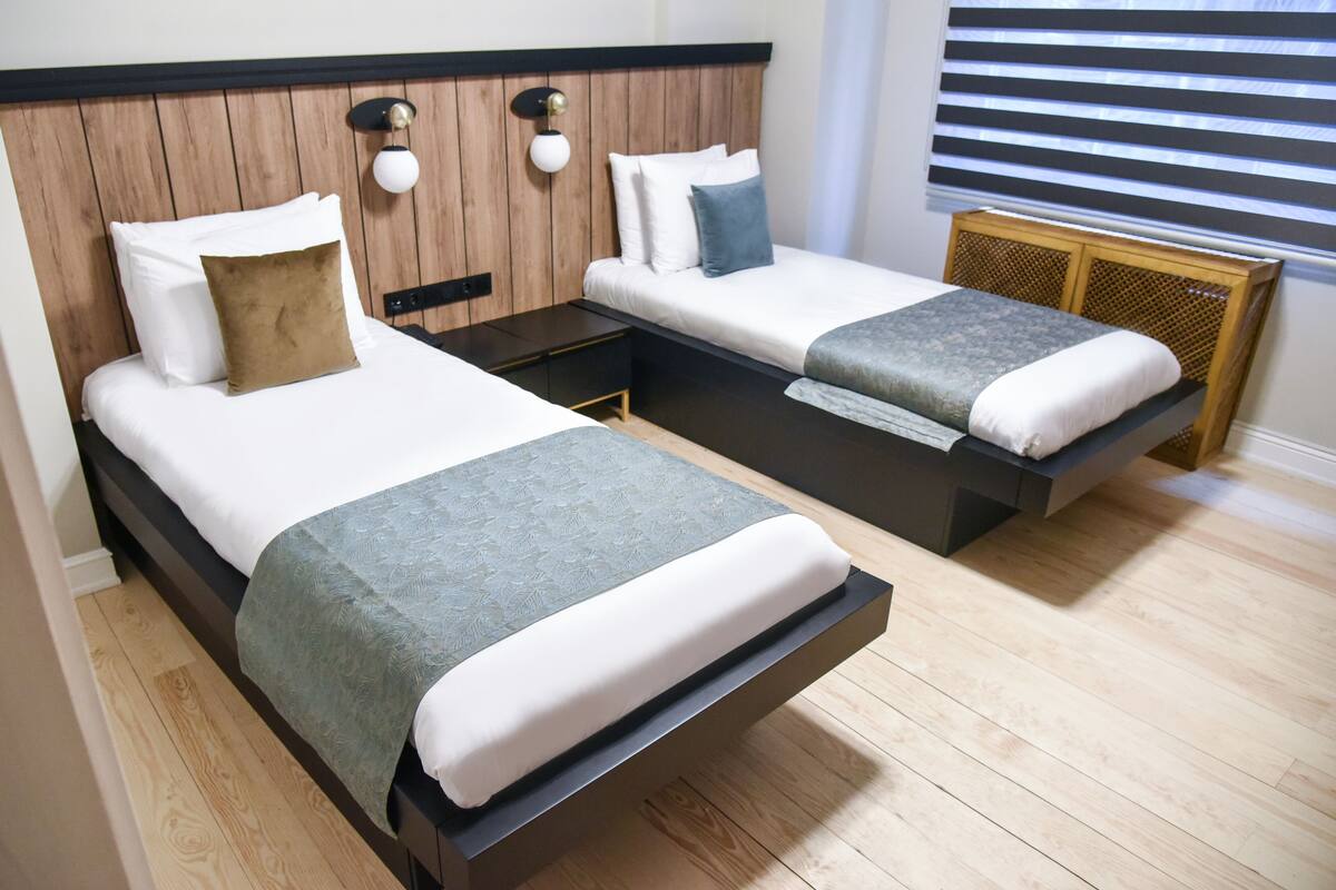 A hotel room with two beds, wooden headboards, white bedding, and a window with blinds.