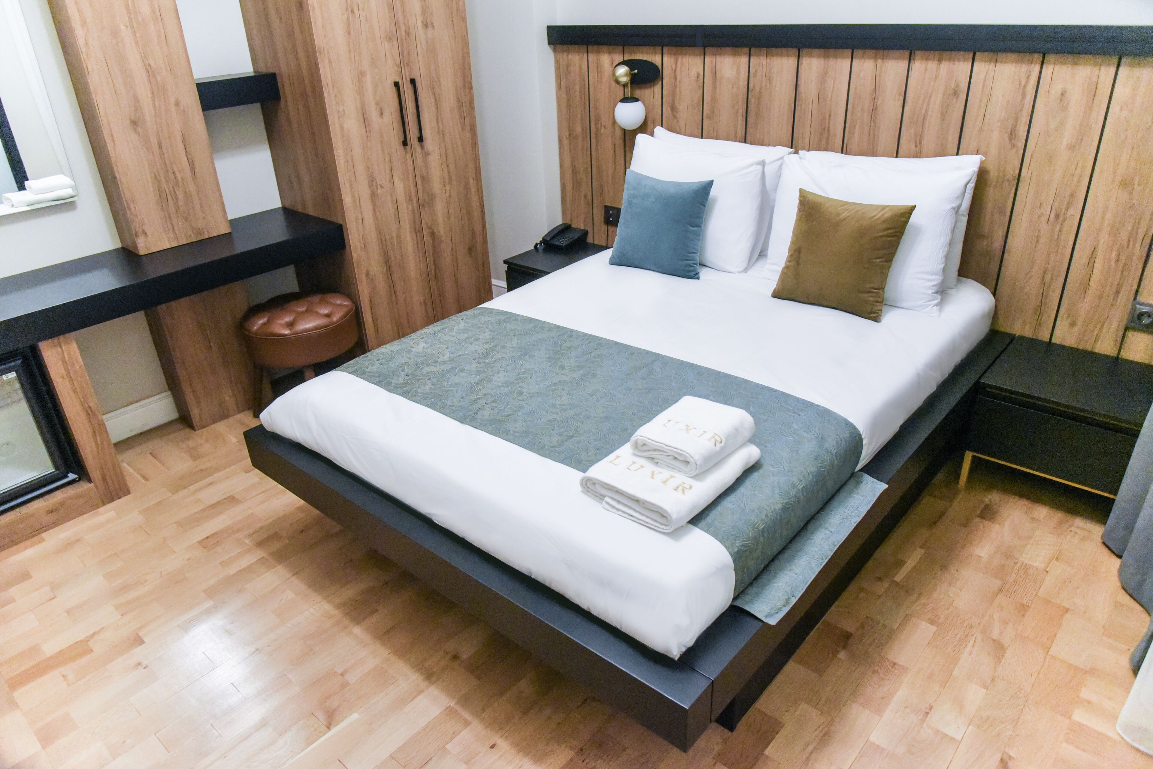 A modern bedroom with a wooden headboard, a large bed with white and blue bedding, a nightstand with a phone, and a wooden bench with a brown cushion.