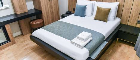 A modern bedroom with a wooden headboard, a large bed with white and blue bedding, a nightstand with a phone, and a wooden bench with a brown cushion.