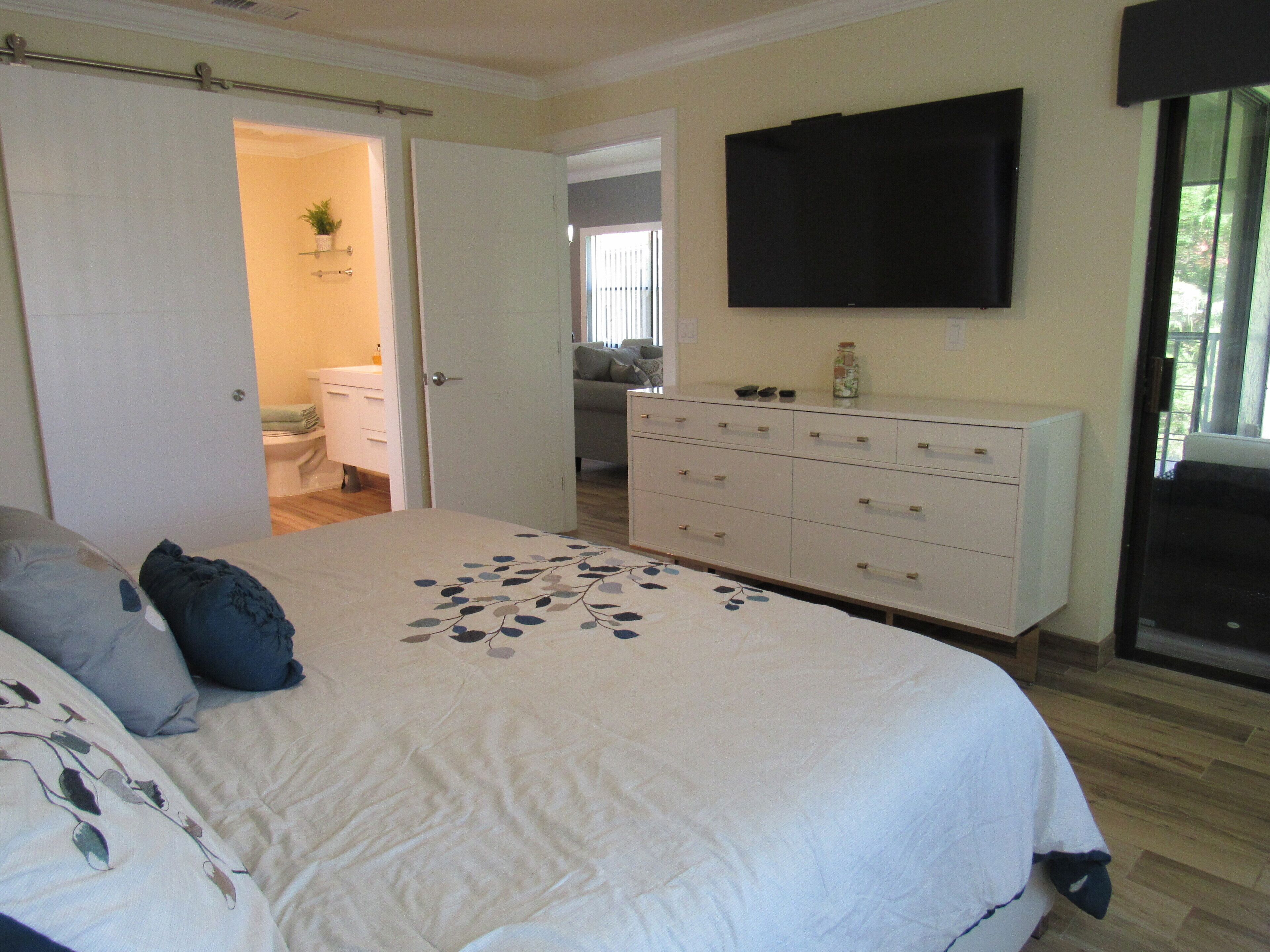 Un dormitorio con cama, tocador, televisor y baño a la vista a través de una puerta abierta.