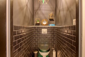 A compact bathroom with a modern design, featuring a toilet, a trash bin, and a small shelf with a plant and a decorative mirror. - La Maison, ★ L'her beau d'hier 3-bedroom house (sleeps 8) 300m from the beach ★ (L'Épine)
