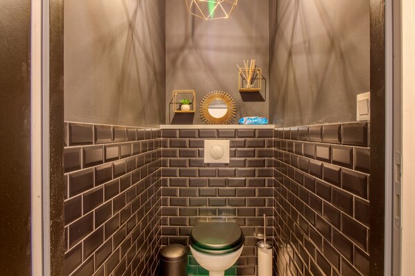 A compact bathroom with a modern design, featuring a toilet, a trash bin, and a small shelf with a plant and a decorative mirror. - La Maison, ★ L'her beau d'hier 3-bedroom house (sleeps 8) 300m from the beach ★ (L'Épine)