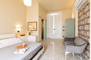 Habitación de hotel con cama, una silla, una estantería y una puerta que da a otra habitación.