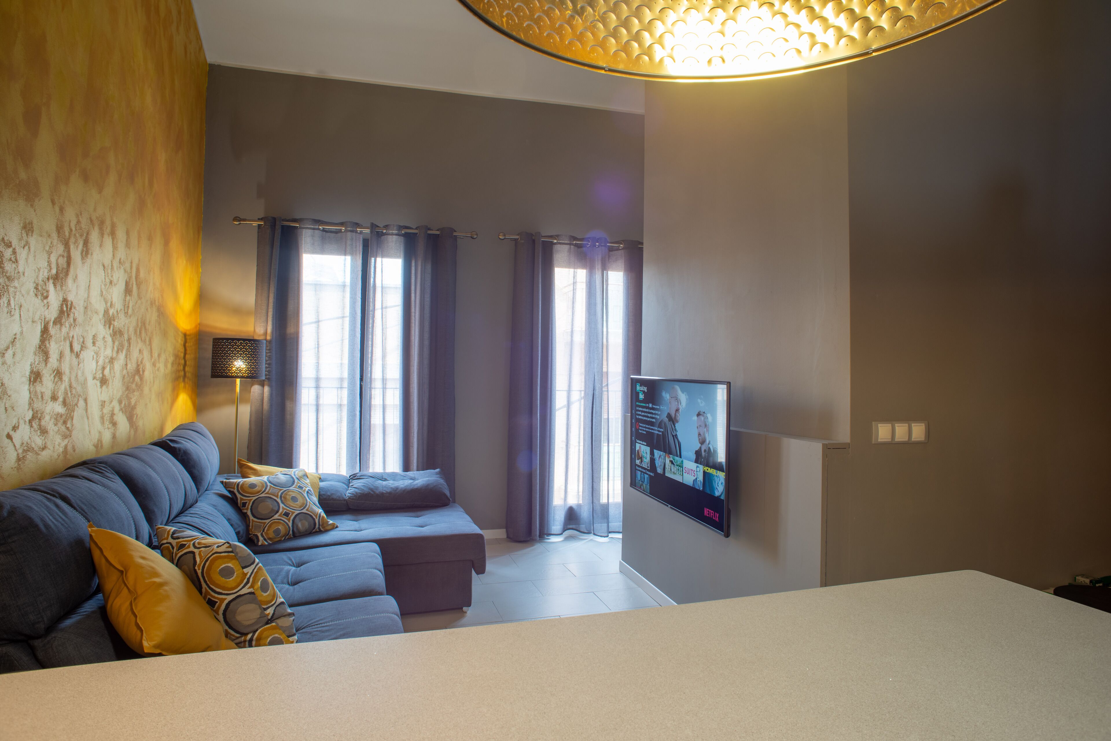 Una sala de estar moderna con un sofá, un televisor de pantalla plana montado en la pared y un ventanal con cortinas.