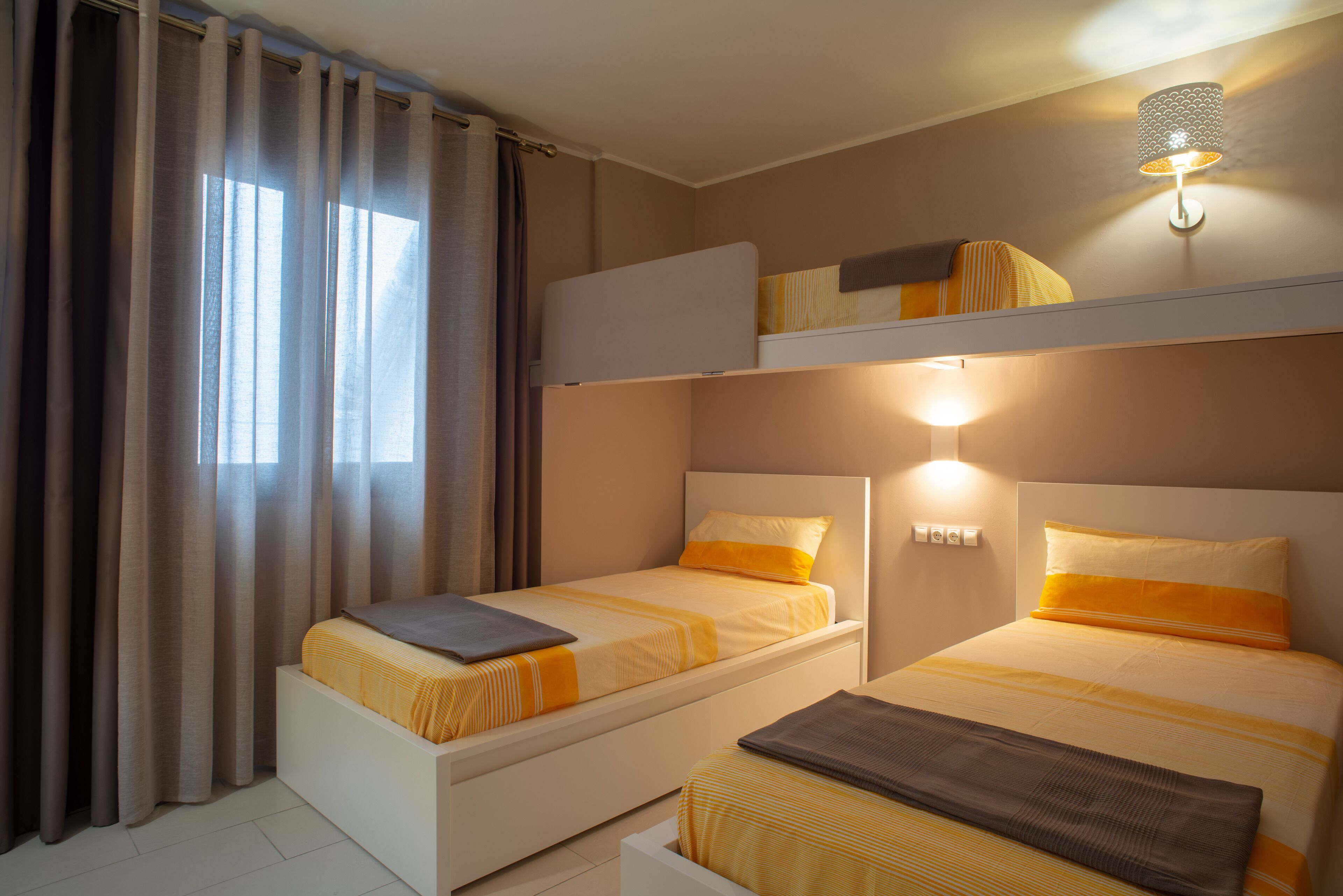 A hotel room with two beds, a bunk bed, a window with curtains, and a wall-mounted light fixture.