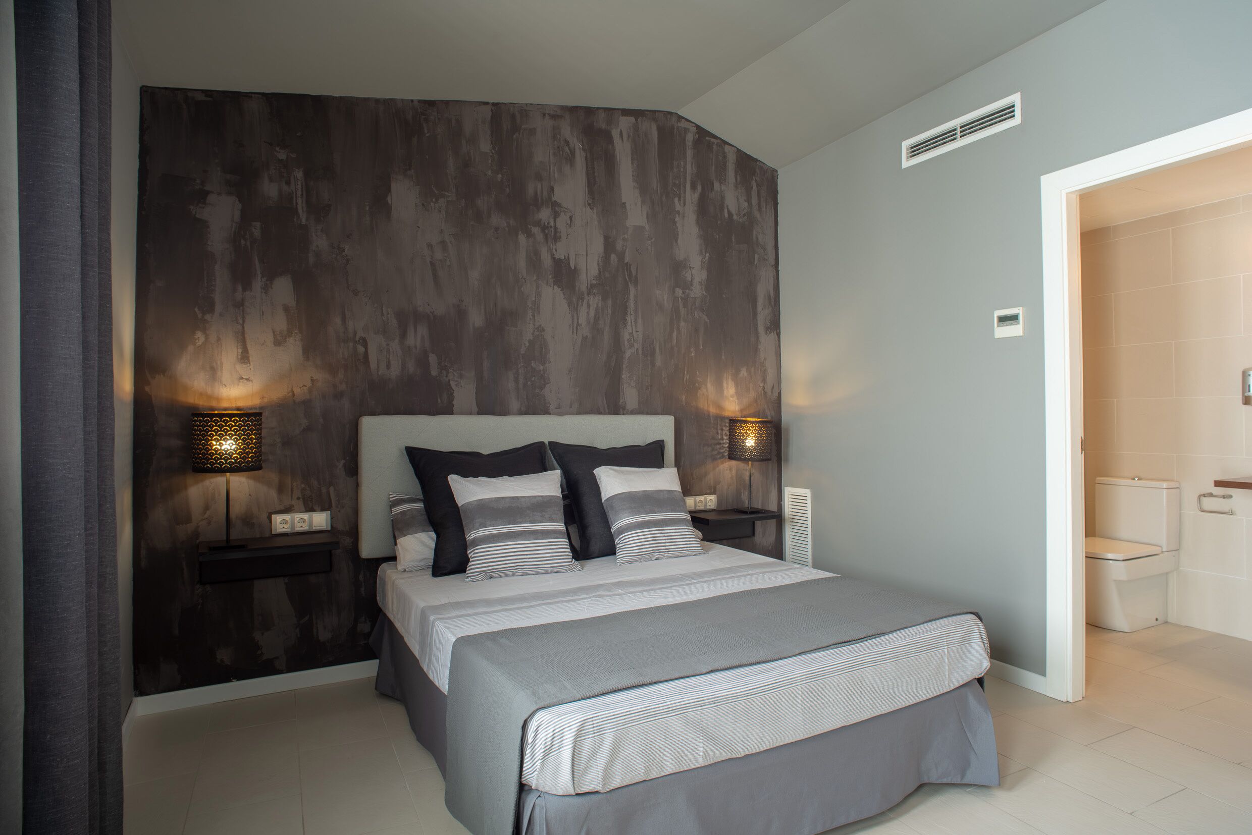 A modern bedroom with a large bed, two bedside tables with lamps, a dark textured wall, and a bathroom visible through an open door.