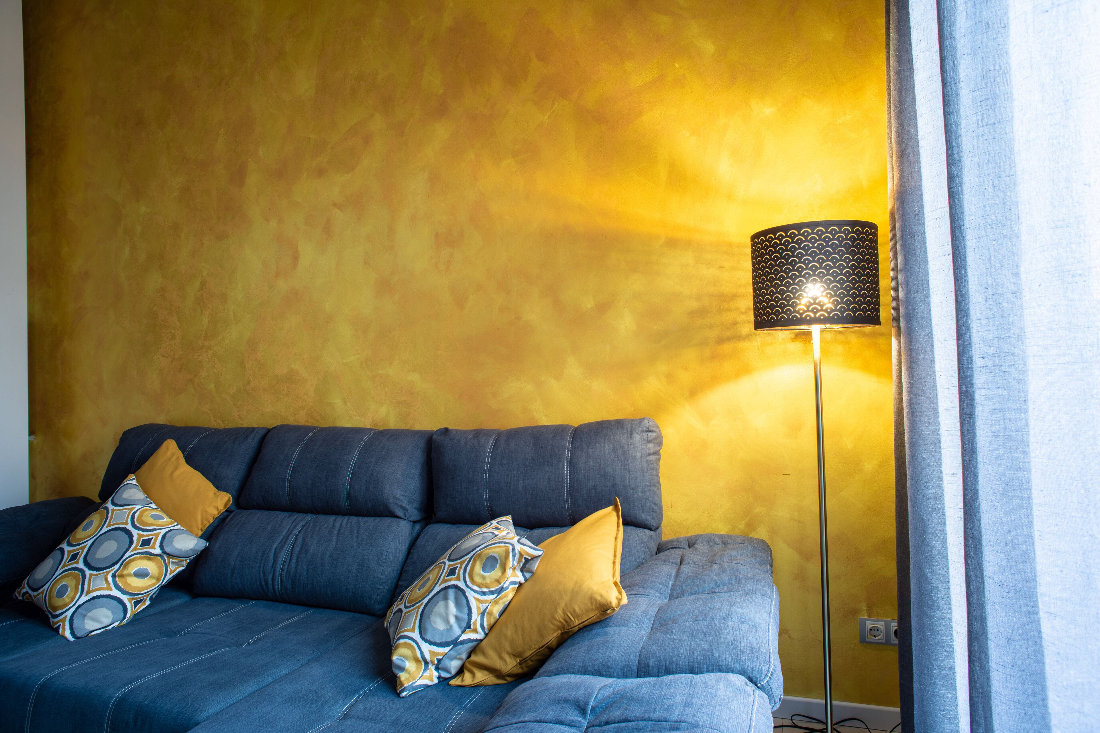 A room with a blue sofa, two patterned pillows, and a floor lamp against a yellow wall.