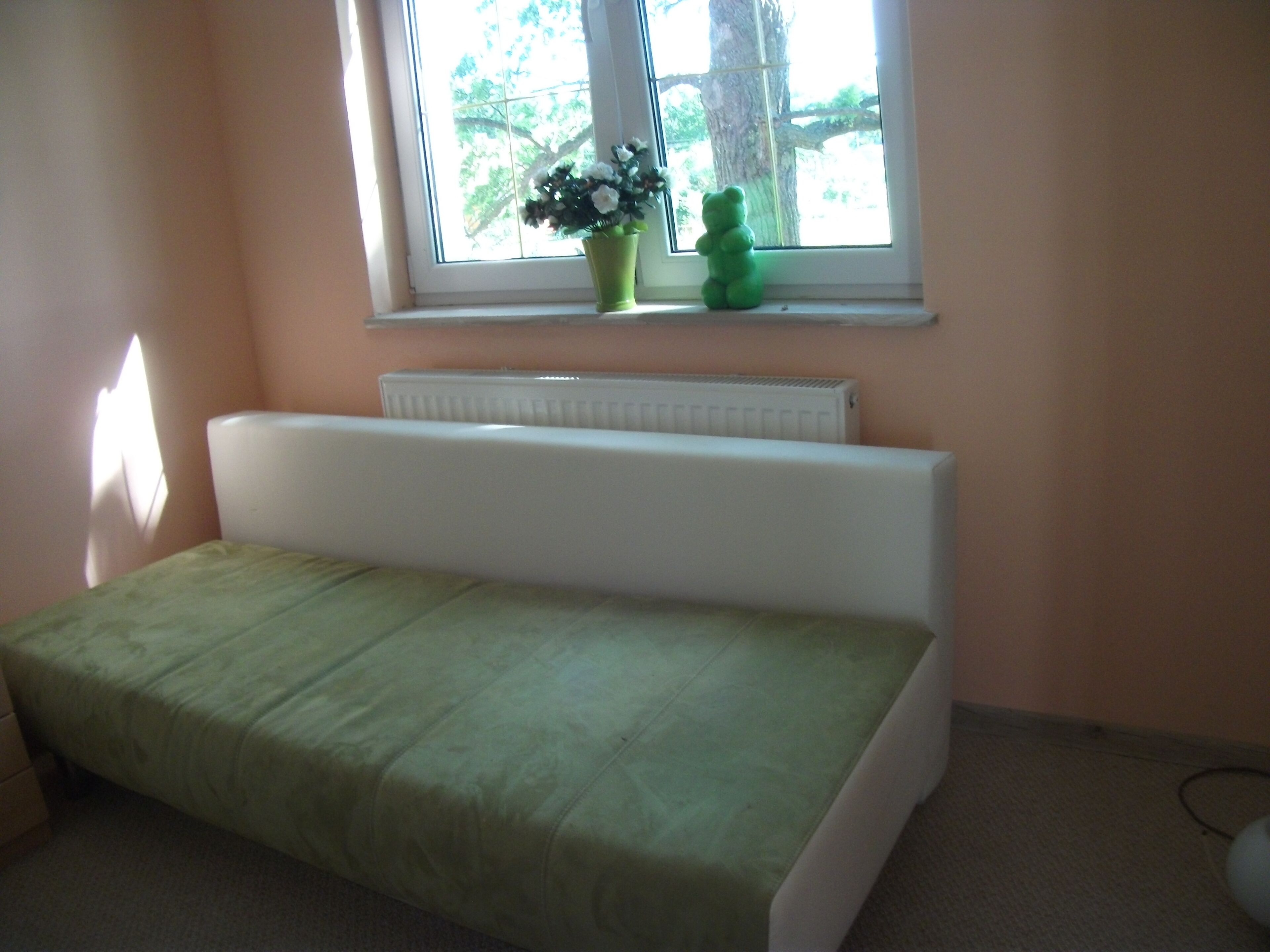 Ein Zimmer mit einem Bett, einem Heizkörper, einem Fenster mit Blick auf Bäume und einem grünen Teddybären auf dem Fensterbrett.