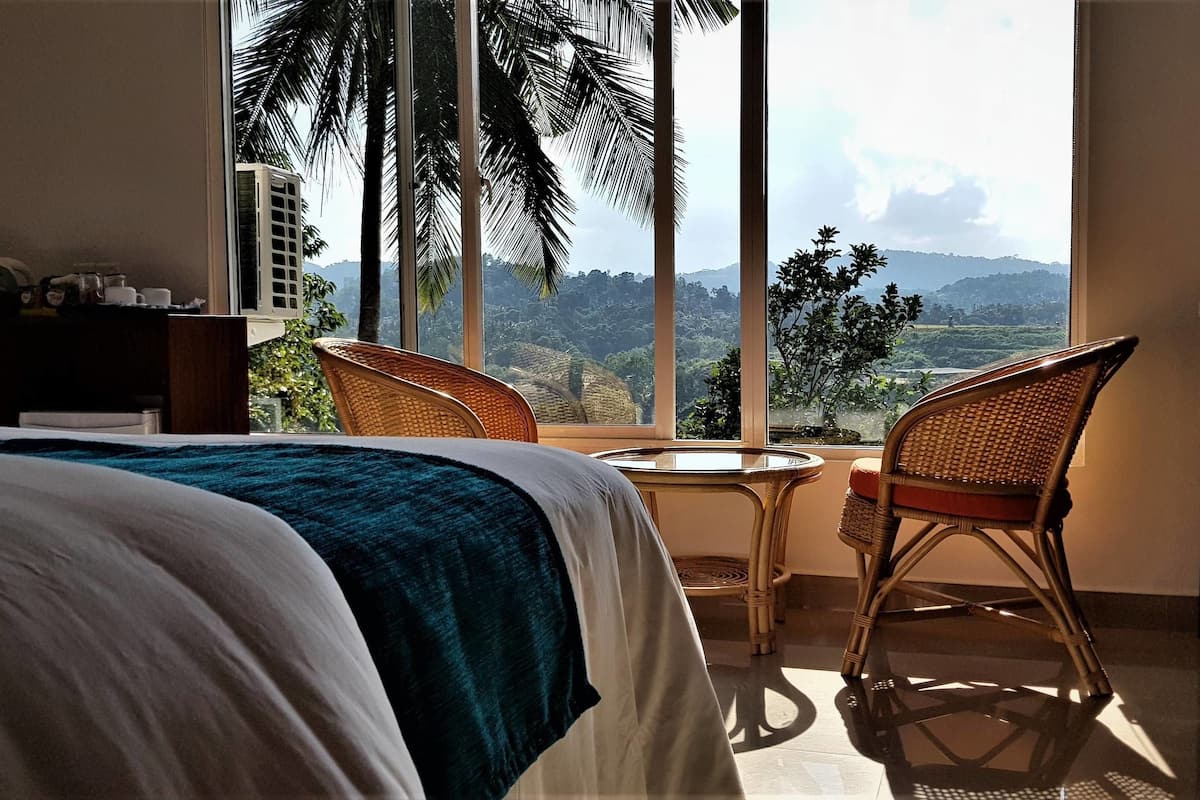 Quarto de hotel com uma cama grande, uma mesa pequena, duas cadeiras de vime e vista para uma paisagem montanhosa através de amplas janelas.