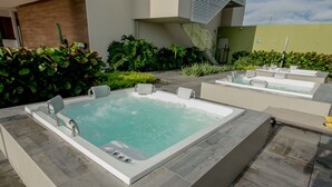 Un área de spa con una bañera grande y otras más pequeñas, rodeada de follaje y arquitectura moderna.