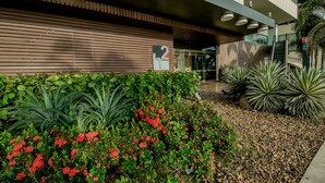 Un edificio moderno con fachada de madera, entrada de vidrio y un jardín ajardinado con flores rojas y plantas de yuca.