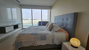Un dormitorio moderno con una cama grande, un cabecero gris y vistas a la ciudad a través de amplios ventanales.