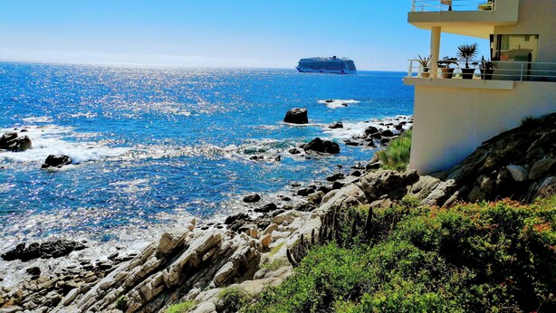 Un hotel sul lungomare con un balcone vista mare, costa rocciosa e una nave da crociera all'orizzonte.