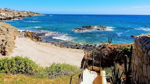 Paesaggio costiero con spiaggia rocciosa, mare blu cristallino e una scalinata che conduce alla riva.