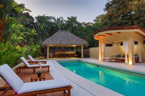A pool area with lounge chairs, a thatched-roof structure, and a covered seating area with curtains. - Luxury Villas of Rancho Manuel Antonio (Quepos)