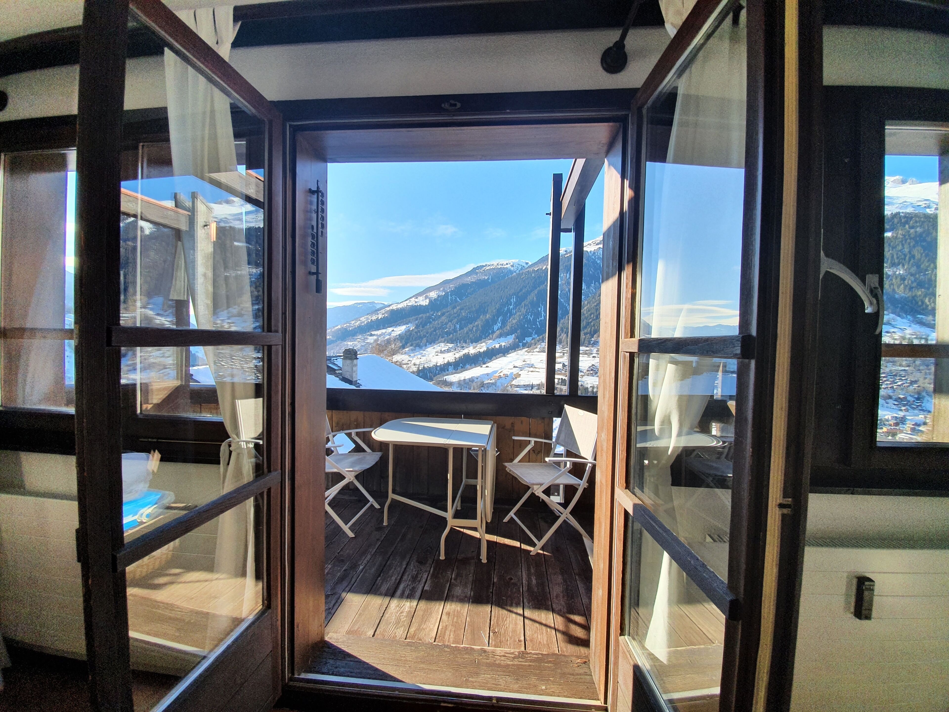 Une terrasse en bois avec une table et des chaises, donnant sur un paysage montagneux enneigé grâce à de grandes fenêtres.