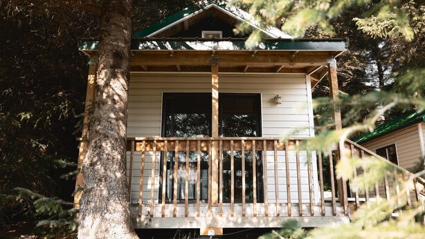 A two-story wooden cabin with a balcony, surrounded by trees. - Cabin 3 Lynn View Lodge (Haines)