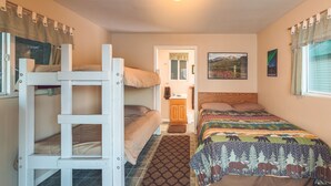A dorm-style room with bunk beds, a small kitchenette, a window with curtains, and a bed with a patterned comforter. - Cabin 3 Lynn View Lodge (Haines)