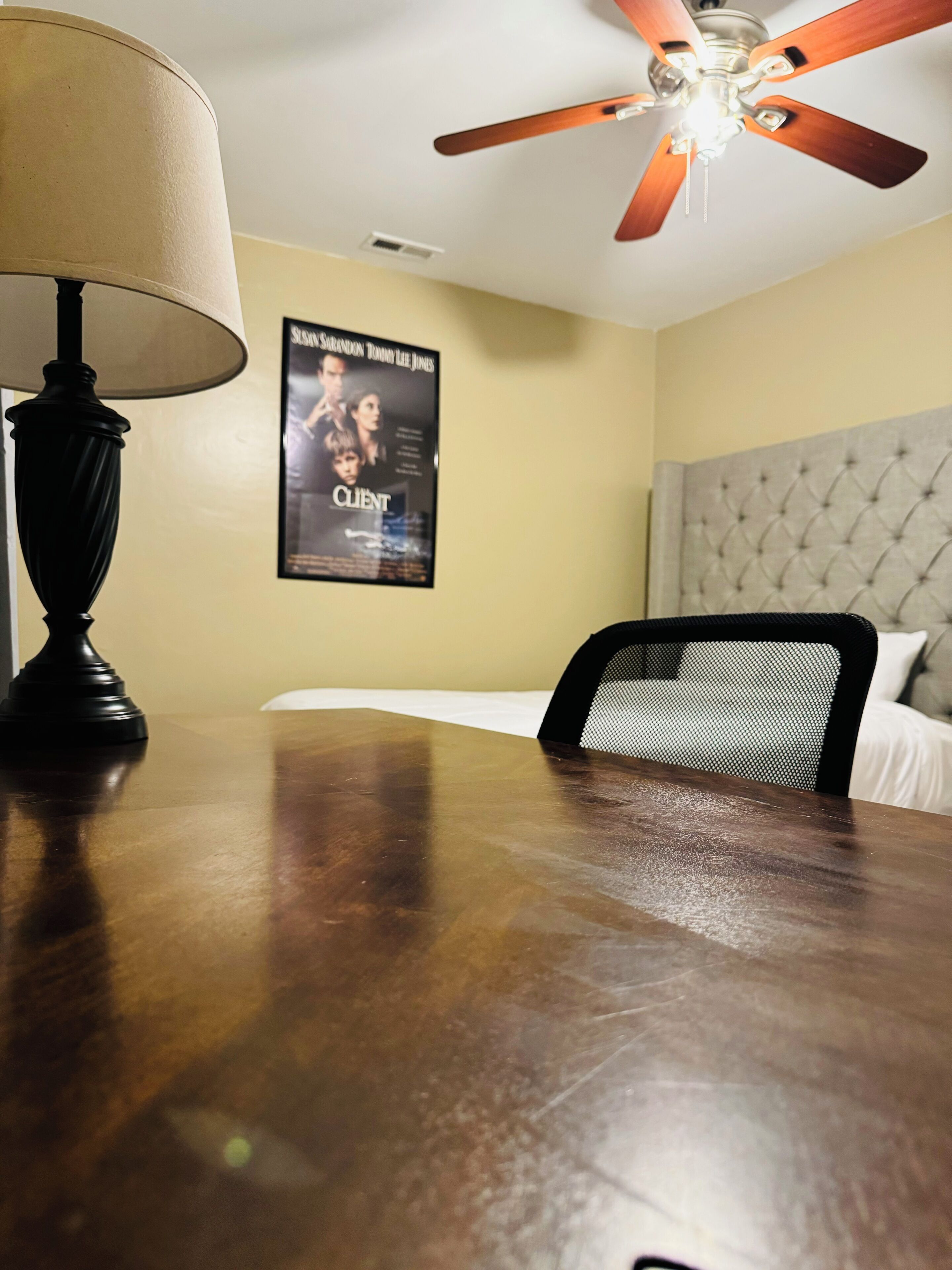 A hotel room with a wooden table, a ceiling fan, a bed with a tufted headboard, and a poster on the wall.