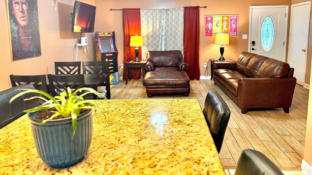 A living room with a brown leather sofa, a dining table with chairs, a ceiling fan, a TV mounted on the wall, and a small arcade game. - NC Music Factory Ranch Getaway (Charlotte)