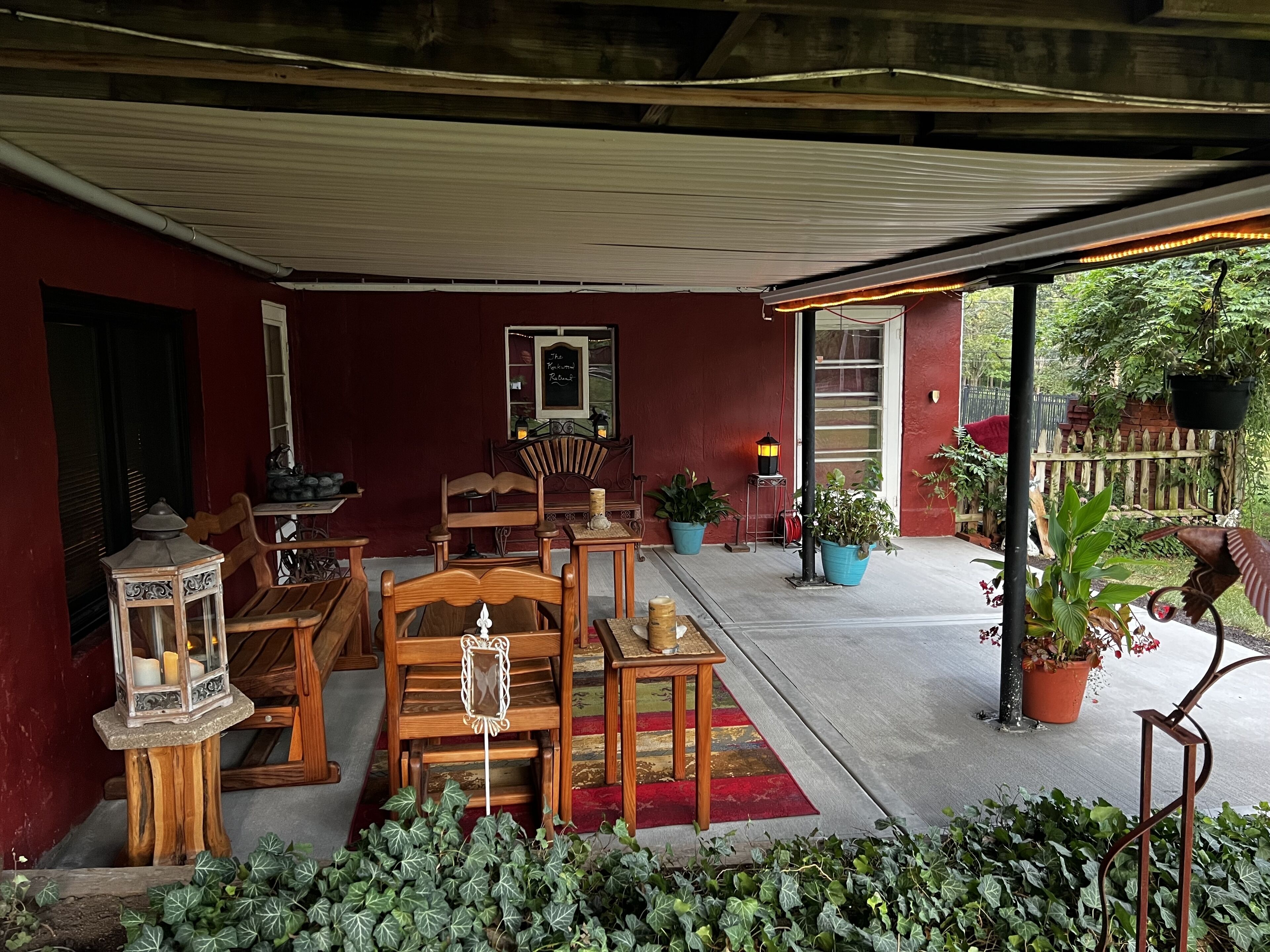 A covered outdoor seating area with wooden furniture, potted plants, and a red wall.
