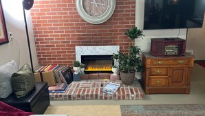 Een woonkamer met een open haard van baksteen, een klok, een televisie en een houten kast.