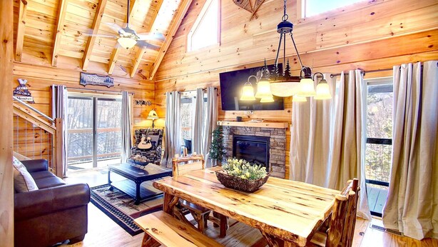 Ein gemütliches Blockhaus-Interieur mit einem Holzesstisch, Bänken, einem Kamin und einem großen Fenster mit Blick ins Freie.