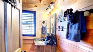Innenansicht einer Holzhütte mit einer Holztür, auf der 'OUTHOUSE' steht, einem Fenster mit Jalousien, einem Waschbecken mit Handtuch und einem Spiegel mit Handtuchhalter.