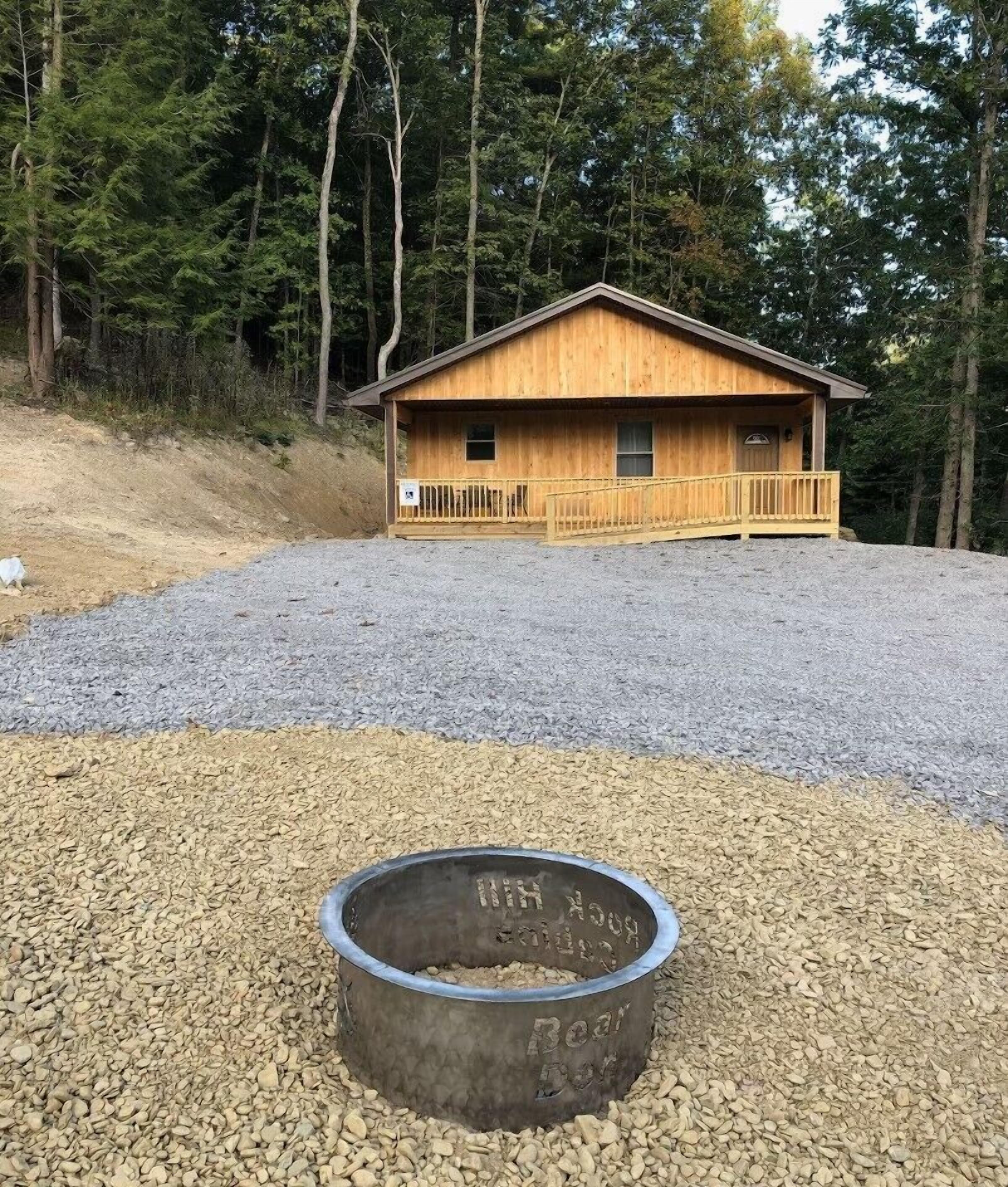 Une cabane en bois dotée d’un porche, entourée de gravier et d’arbres.
