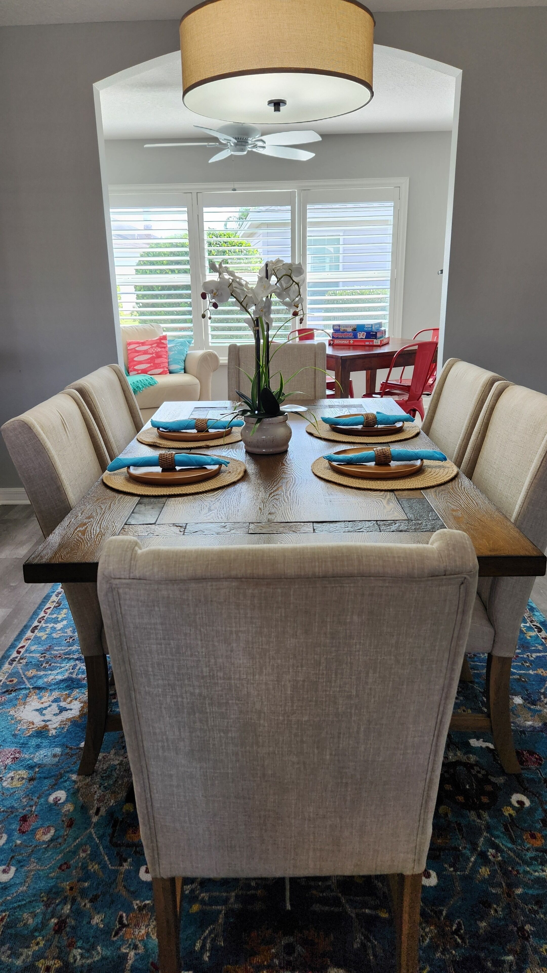 A dining area with a wooden table set for six, surrounded by upholstered chairs. A ceiling fan and a large window with blinds are visible in the background.