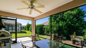 A patio with a glass table and chairs, a ceiling fan, and a view of a backyard with a lawn and trees. - Ruhe Haus in Collier. 3/2, FREE golf, 4-seat cart, RESIDENT passes, pets (The Villages)