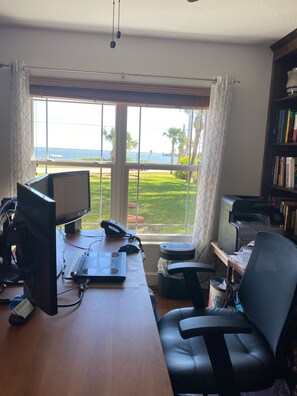 Escritório em casa com mesa, computador, impressora e cadeira. Uma janela oferece vista para uma área gramada e um corpo d'água.