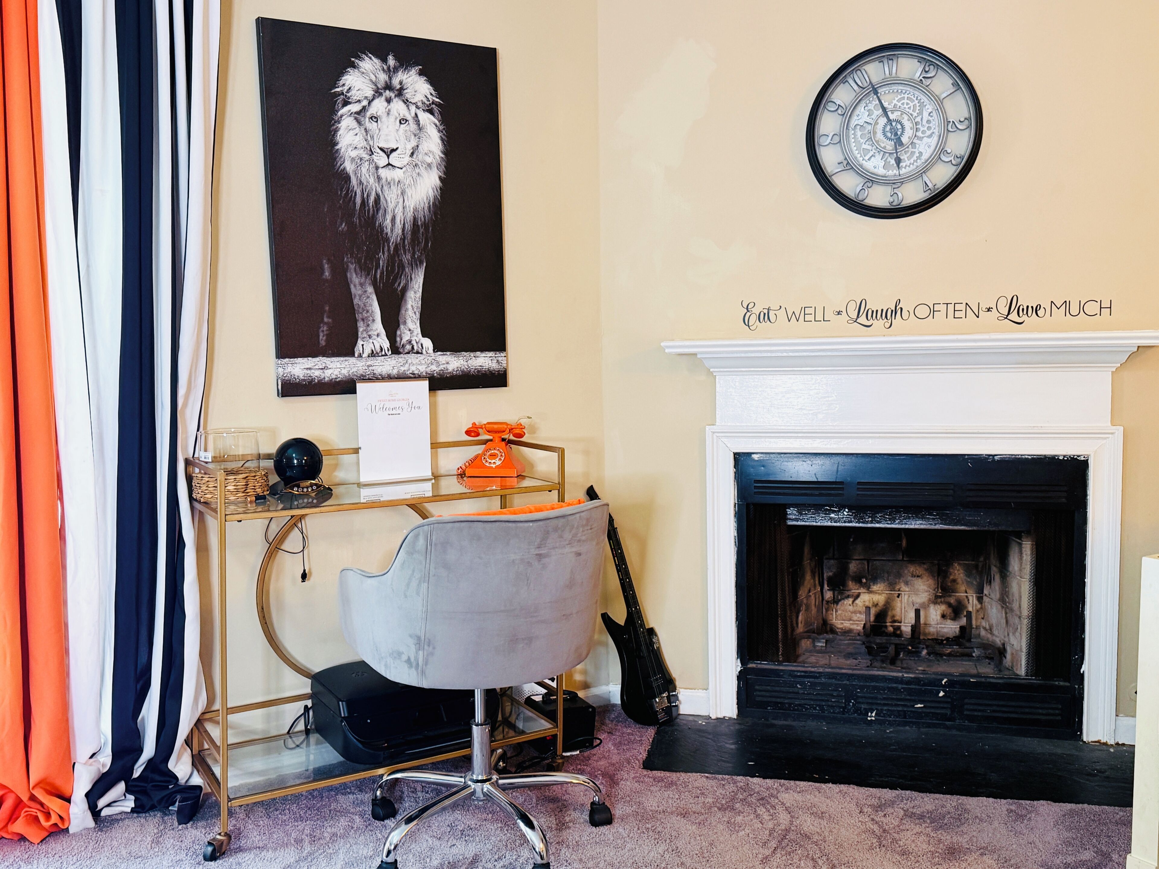 A room with a fireplace, a clock, a desk with a chair, a framed picture of a lion, and a sign above the fireplace.