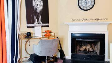 A room with a fireplace, a clock, a desk with a chair, a framed picture of a lion, and a sign above the fireplace.