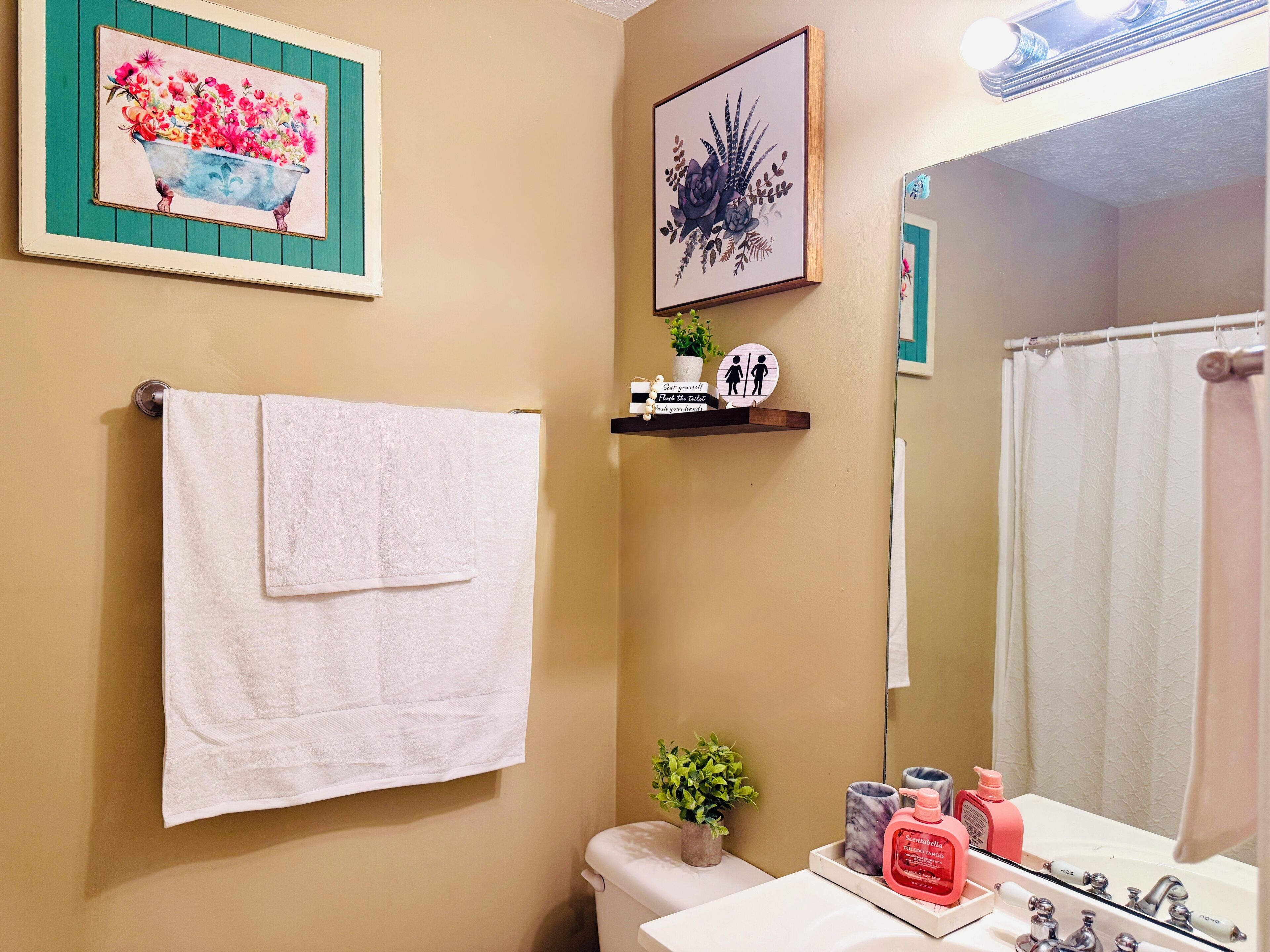 Une salle de bain avec une serviette blanche suspendue à un porte-serviettes mural, une étagère contenant des articles de toilette, une œuvre d’art florale encadrée et un miroir qui reflète le rideau de douche.